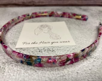 May include: A thin, circular headband featuring a multicoloured tortoiseshell pattern with shades of pink, white, blue, and brown. A white square card is positioned inside the headband, displaying a line drawing of a person and the text For the Hair you wear.
