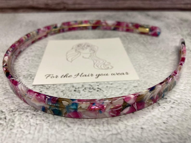 May include: A thin, circular headband featuring a multi-colored, marbled pattern with shades of magenta, white, blue, and brown. The headband is positioned around a square white card that displays a line drawing of a person and the text For the Hair you wear.