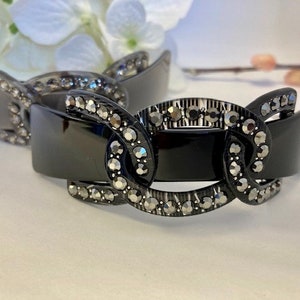 Peut inclure: Une pince à cheveux noire avec un motif décoratif de cercles entrelacés ornés de strass.