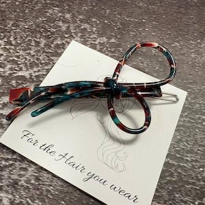May include: A bow-shaped hair clip with a marbled pattern in shades of red, teal, and white. The clip is made of a shiny material and is resting on a white card with the text "For the Hair you wear".