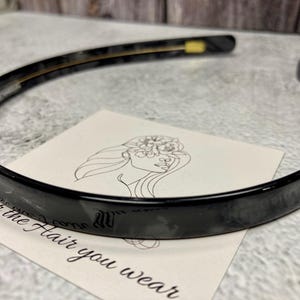 May include: A glossy black plastic headband with a thin, smooth, and curved design. The headband features small gold-colored metal accents on the inner sides. It is positioned over a white card with a line drawing of a face and the text For the Hair you wear.