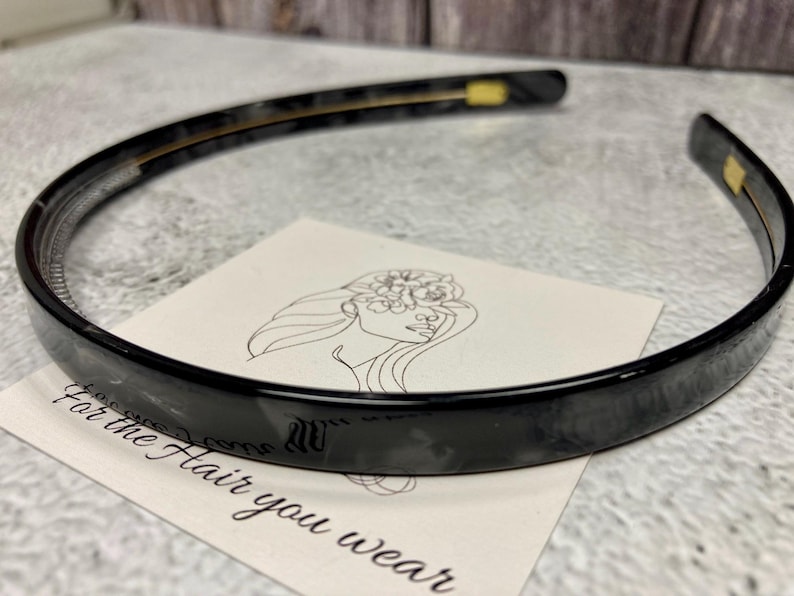 May include: A glossy black plastic headband with a thin, smooth, and curved design. The headband features small gold-colored metal accents on the inner sides. It is positioned over a white card with a line drawing of a face and the text For the Hair you wear.