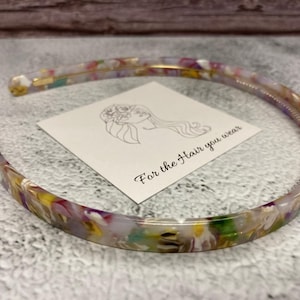 May include: A thin, semi-circular headband made of multi-colored, marbled acrylic material featuring shades of yellow, purple, green, and white. A square white card is placed inside the headband with a line drawing of a person and the text For the Hair you wear.
