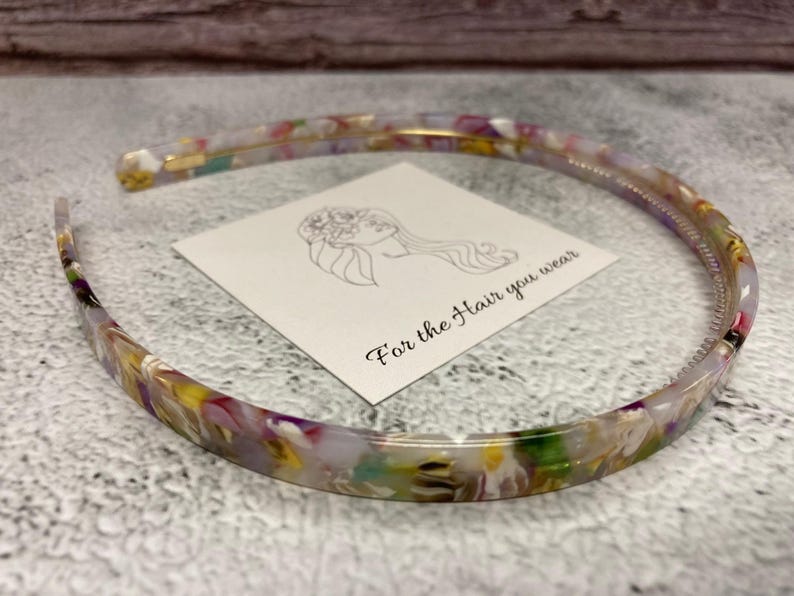 May include: A thin, semi-circular headband made of multi-colored, marbled acrylic material featuring shades of yellow, purple, green, and white. A square white card is placed inside the headband with a line drawing of a person and the text For the Hair you wear.