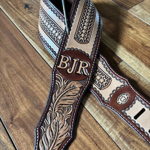 May include: A handcrafted leather guitar strap with a detailed feather design and the initials "BJR" embossed on the strap. The strap features a light brown and dark brown color scheme with decorative stitching and a metal hook.