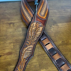May include: A brown leather guitar strap with intricate carved feather and floral designs. The strap features a basket weave pattern and adjustable slots for length. The edges are stitched with white thread, and the strap has a metal attachment.