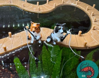 以下が含まれることがあります： 茶色と白のミニチュア犬と黒と白のミニチュア犬の2匹が、木製の桟橋に座って池で釣りをしています。犬たちは釣り竿を持っており、糸を水中に垂らしています。池は緑の植物に囲まれています。