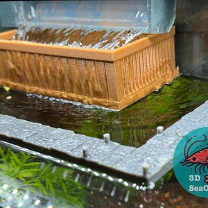 May include: A miniature aquarium decoration featuring a brown wooden-style waterfall and a grey stone walkway. The water flows from a clear container into the miniature pond. A 3D SeaCrafts logo is visible.
