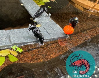 以下が含まれることがあります： 灰色の石の道に3羽の黒いカラスの置物が置かれ、1羽がオレンジ色のボールを持っています。道は水と緑の植物に囲まれています。画像には3D SeaCraftsのロゴが含まれています。