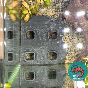 以下が含まれることがあります： 複数の正方形の開口部のある黒いプラスチック製の水槽の背景。背景は、小さな魚やエビの隠れ場所を提供するように設計されています。背景は、水と砂利に部分的に浸かっています。