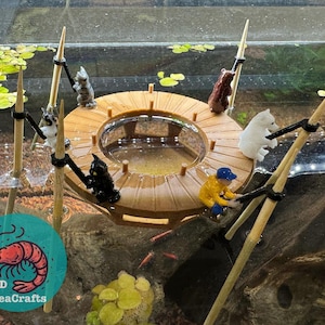 May include: A wooden platform with a clear glass bowl in the center, surrounded by small figurines of animals and people. The platform is attached to several wooden sticks that are sticking up from the bottom of the platform. The platform is in a fish tank with water and plants.