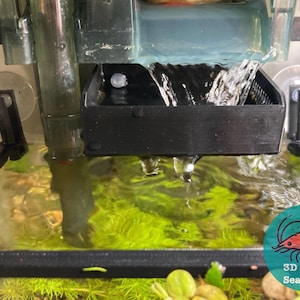 May include: A black plastic filter for an aquarium with a clear plastic tube and a water flow cascading into a clear glass tank with green plants and a red shrimp logo on the bottom right corner that says "3D SeaCrafts".