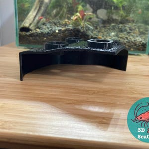 May include: Black 3D printed aquarium plant holder with three openings. The holder is curved and has a rounded edge. The holder is sitting on a light brown wooden surface. 3D SeaCrafts logo is visible in the lower right corner.