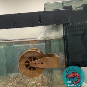 Puede incluir: Un alimentador de acuario de plástico marrón con una rueda y un pequeño contenedor. El alimentador está unido al lado de un acuario de vidrio. El alimentador está diseñado para dispensar comida para peces automáticamente.