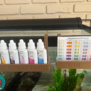 Puede incluir: Un soporte de plástico marrón con seis botellas transparentes y una tarjeta blanca con el texto "Freshwater Master Test Kit" y tablas de colores para probar los parámetros del agua. El soporte está unido al lado de un acuario de vidrio.