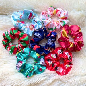 May include: A set of eight colorful Christmas-themed scrunchies. The scrunchies are made of satin and feature various holiday designs, including Santa Claus, snowmen, and candy canes.