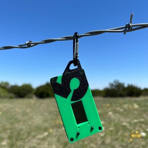 May include: A green and black keychain with a circular design hangs from a barbed wire fence. The keychain has a small screen and a few buttons.