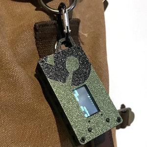 May include: A small, green, rectangular device with a black screen and a black and white design on the front. The device is attached to a brown leather strap with a metal ring.