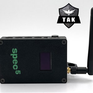 May include: Black rectangular device with a screen, antenna, and the text "spec5" in green. The device has a button on top and is labeled "TAK" with a shield logo. The antenna is black and adjustable.