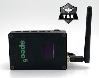 May include: Black rectangular device with a screen, antenna, and the text "spec5" in green. The device has a button on top and is labelled "TAK" with a shield logo. The antenna is black and adjustable.