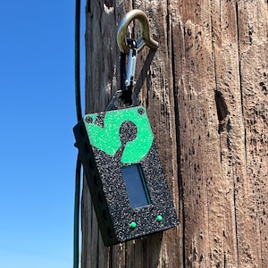 May include: A black and green device with a small screen and a green logo attached to a wooden pole with a metal clip.