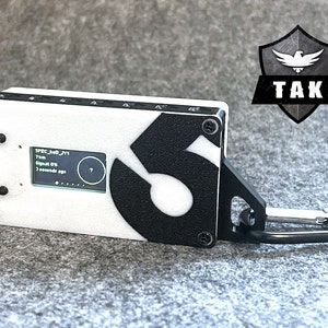 May include: A white and black device with a small screen displaying "SPEC_had_JY1 7km Signal 0% 7 seconds ago". The device has a black number "6" on the side and a black carabiner clip.