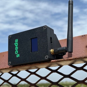 May include: A black device with a small screen and a green "spec5" label attached to a metal fence. The device has a black antenna extending from the top.