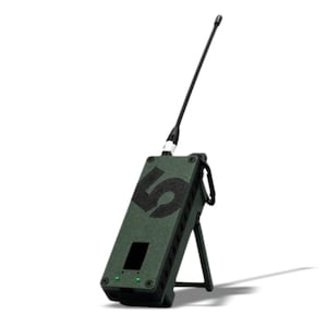 May include: An olive green electronic device with a black antenna and the number "5" printed on the front. The device has a small screen and two green lights. It is propped up by a black stand and has a black carabiner attached.