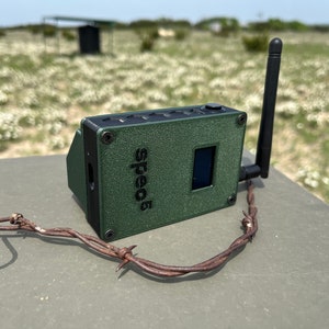 May include: A green, textured, rectangular device with a black antenna and a small screen. The device has the word "speed5" printed on it. The device is sitting on a gray surface with a rusty barbed wire.