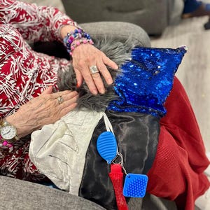 May include: A blue sequined pillow, a white satin fabric, and a red corduroy fabric are visible. A blue and red keychain with a textured design hangs from the fabrics. A person's hands and arms are also visible, adorned with jewelry.