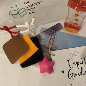 Puede incluir: Una pelota antiestrés de color rosa en forma de estrella con el texto "You are amazing!" en ella, una bolsa de cuero marrón, una bolsa naranja, una bolsa de pelo negro, un juguete magnético rojo y blanco, una caja de plástico transparente con el texto "The Cognitive Busy Box", una postal con una imagen del Puente Golden Gate y un reloj de arena de plástico transparente con líquido rojo y amarillo.