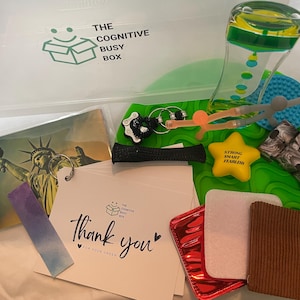 May include: A clear plastic storage container filled with various items, including a green and yellow hourglass, a blue and green sensory toy, a star-shaped stress ball with the words "Strong Smart Fearless", a black and white fidget toy, a postcard with a picture of the Statue of Liberty, a black headband, a set of silver keychains, a red purse, a white thank you card, and a brown fabric square.