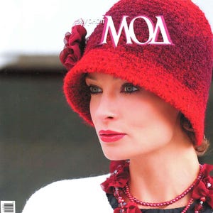 May include: A red knitted hat with the word "MODA" embroidered on the front. The hat is worn by a woman with a red necklace and a white sweater. The magazine cover reads "ШАПОЧКИ № 561".