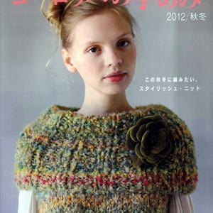 以下が含まれることがあります： カラフルな手編みの襟巻きを着用した女性。大きな濃い緑色の花のアクセントが付いています。襟巻きは緑、赤、黄色の糸で構成されています。背景には日本の編み物雑誌の表紙が見えます。