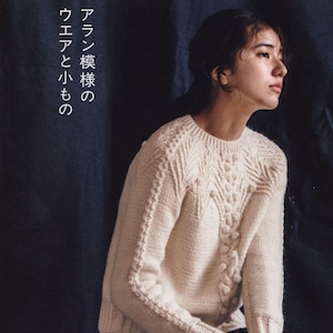 Op de afbeelding: Een crèmekleurige kabelgebreide trui met ingewikkelde patronen en een ronde halslijn. De mouwen en de voorkant hebben gedetailleerde texturen. De afbeelding bevat Japanse tekst: "Aran patronen, kleding en kleine items" en "Let's knit series".