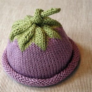 Könnte beinhalten: Eine gestrickte Mütze in Form einer Aubergine. Die Mütze ist hauptsächlich lila mit einer grünen, blattartigen Oberseite. Der Rand ist ein dunklerer Lilaton. Die Mütze ist aus weichem Garn gefertigt, wahrscheinlich für ein Baby oder Kind.