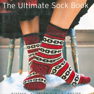May include: The Ultimate Sock Book by Vogue Knitting. The image shows a person wearing red, black, and white patterned socks. The socks are on a light blue chair. The book's title is at the top of the image, with the words "history", "technique", and "design" at the bottom.