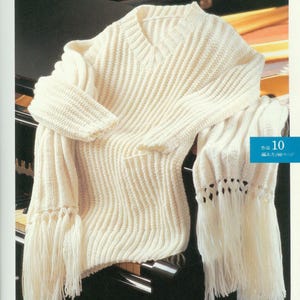May include: A white knitted sweater with a V-neck and long sleeves. The sweater has a fringe trim and is draped over a black piano.