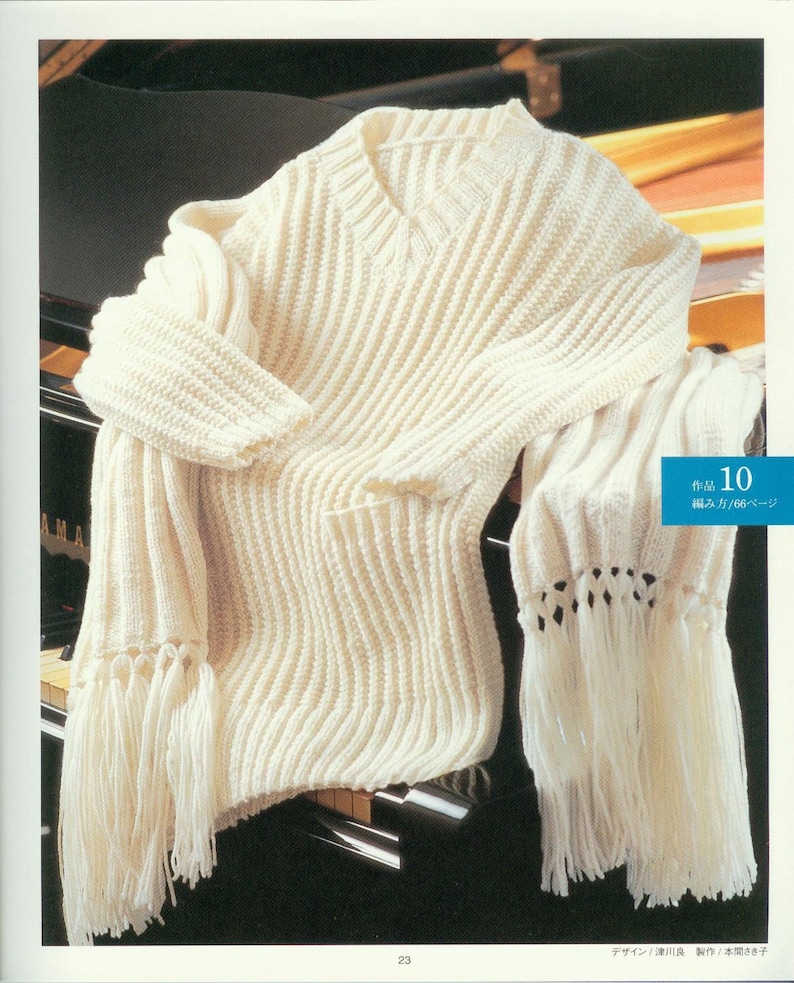 May include: A white knitted sweater with a V-neck and long sleeves. The sweater has a fringe trim and is draped over a black piano.