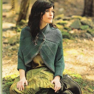 Peut inclure: Une femme portant un cardigan en tricot vert avec un haut en soie marron et une jupe verte. Le cardigan a un design unique avec un motif en losange. La femme est assise dans un décor forestier avec de la mousse et des rochers. Le texte "woodland gothic" est visible en bas de l'image.