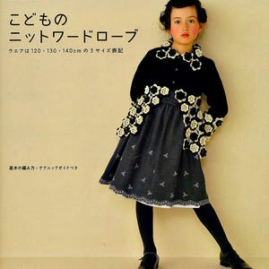 May include: A black and white knit cardigan with a crocheted flower design. The cardigan is worn over a black dress with a white lace trim. The model is a young girl with dark hair and is wearing black tights and black shoes.