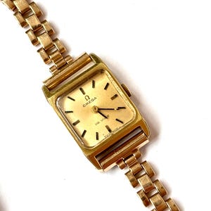 May include: A gold-tone Omega De Ville wristwatch with a rectangular case and a matching link bracelet. The watch face features a gold dial with black hour markers, black hands, and the Omega logo and text printed in black on the dial surface.