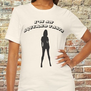 May include: A white t-shirt with a black silhouette of a woman and the text "I'm an acquired taste" printed on the front.