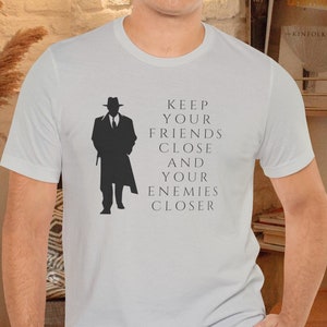Könnte beinhalten: Ein weißes T-Shirt mit einer schwarzen Silhouette eines Mannes in einem Fedora und einem Trenchcoat. Der Text "Keep your friends close and your enemies closer" ist auf das T-Shirt gedruckt.