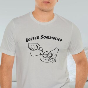May include: A light gray t-shirt with a black graphic of a hand pouring coffee into a cup. The text "Coffee Sommelier" is printed on the shirt.