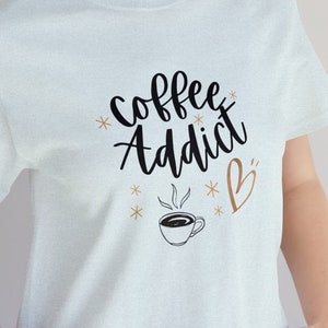 May include: A light blue t-shirt with the text "Coffee Addict" in black cursive lettering. A small coffee cup with steam and a heart are also printed on the shirt.