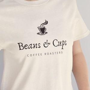 May include: A white t-shirt with a black graphic of a coffee cup with steam and the text "Beans & Cups Coffee Roasters".