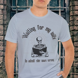 May include: A light blue t-shirt with a skeleton reading a book. The text on the shirt reads "Waiting for my wife to admit she was wrong."