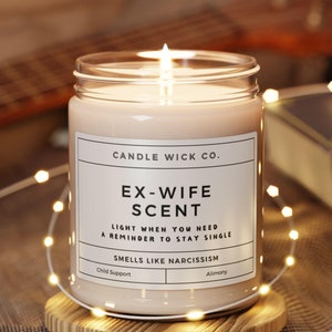 May include: A white candle in a glass jar with a black label that reads "CANDLE WICK CO. EX-WIFE SCENT LIGHT WHEN YOU NEED A REMINDER TO STAY SINGLE SMELLS LIKE NARCISSISM Child Support Alimony".