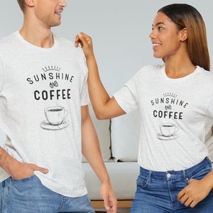 May include: Two people wearing white t-shirts with a black graphic that says "Sunshine and Coffee" and a drawing of a coffee cup. The person on the left is wearing blue jeans. The person on the right is wearing blue jeans and is touching the person on the left's shoulder.